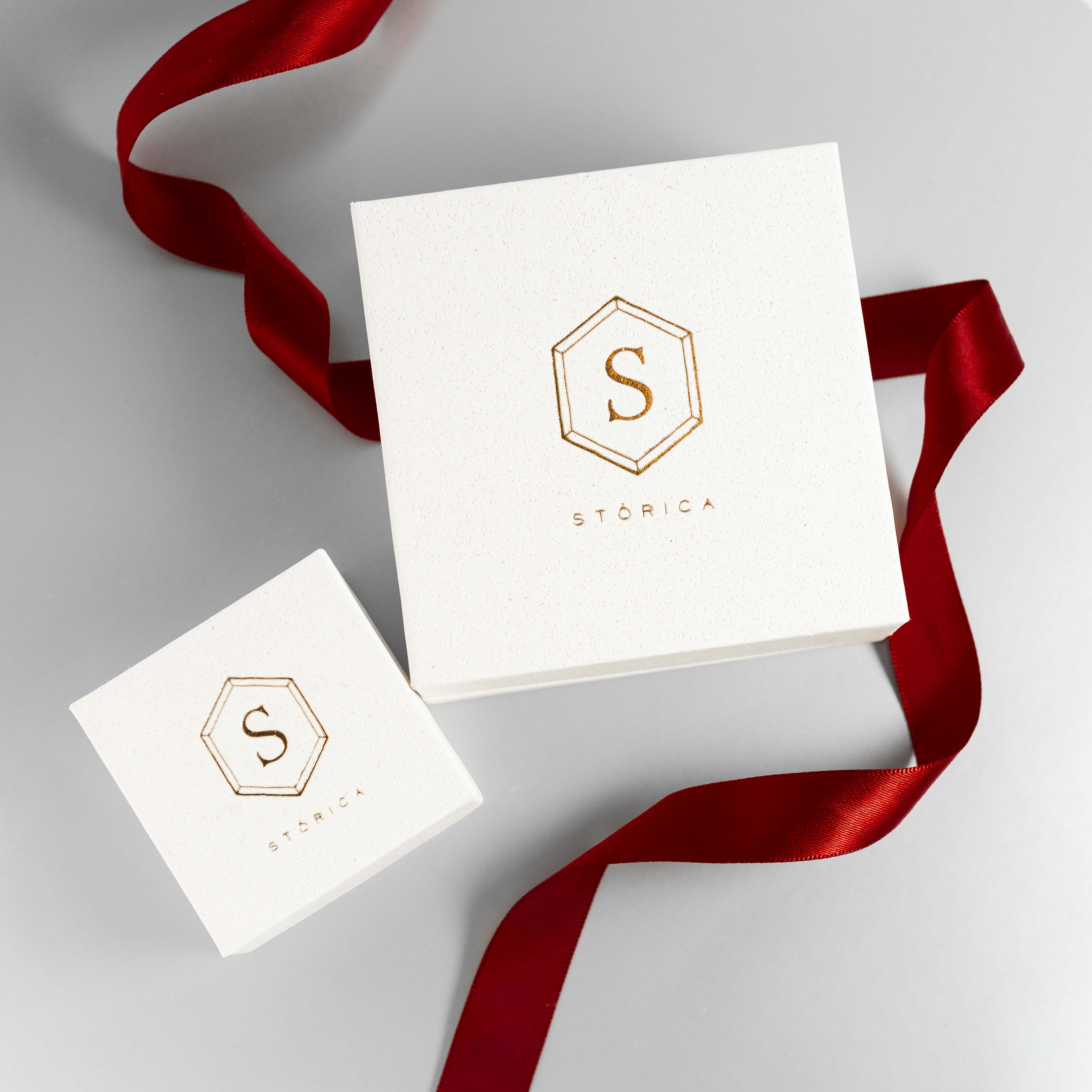 A white box with a gold hexagonal logo on it, tied with a red ribbon, potentially representing the packaging for a piece of jewelry from Stórica.